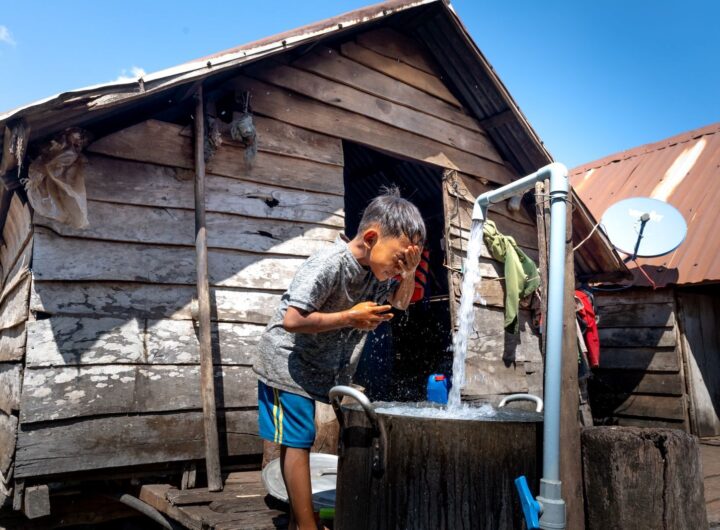 A young boy washes his face at an outdoor tap beside a wooden hut in a rural area.