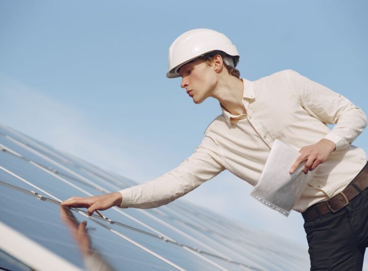 Low angle of young male inspector with documents in formal wear and hardhat standing against solar panels and checking details of construction