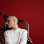 Thoughtful young bald female with disability in warm sweater leaning on wheelchair and looking away against red background