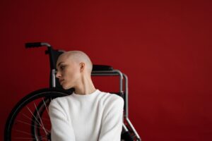 Thoughtful young bald female with disability in warm sweater leaning on wheelchair and looking away against red background