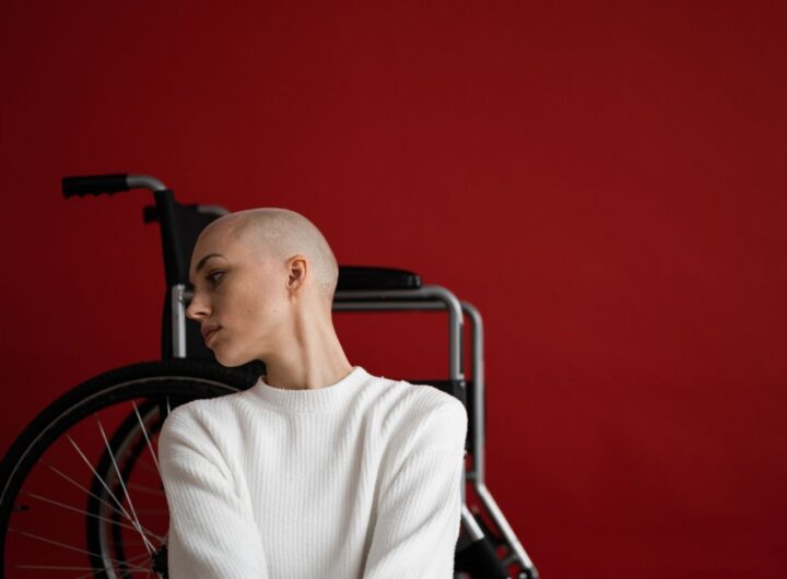Thoughtful young bald female with disability in warm sweater leaning on wheelchair and looking away against red background