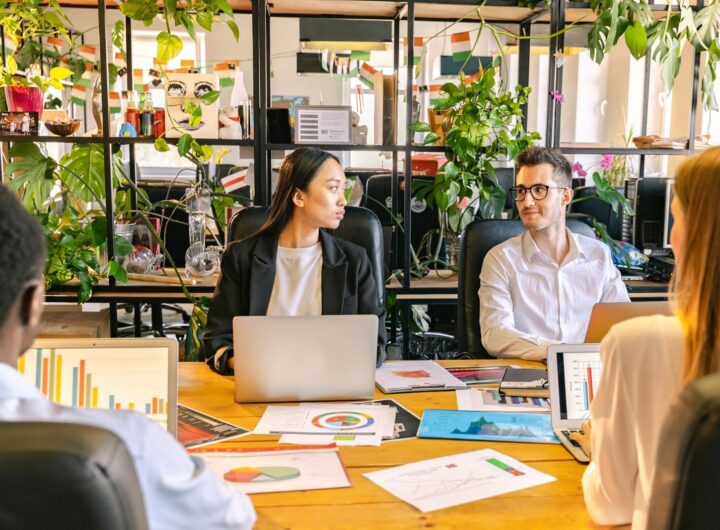 A diverse team of professionals discussing projects in a plant-filled office setting.