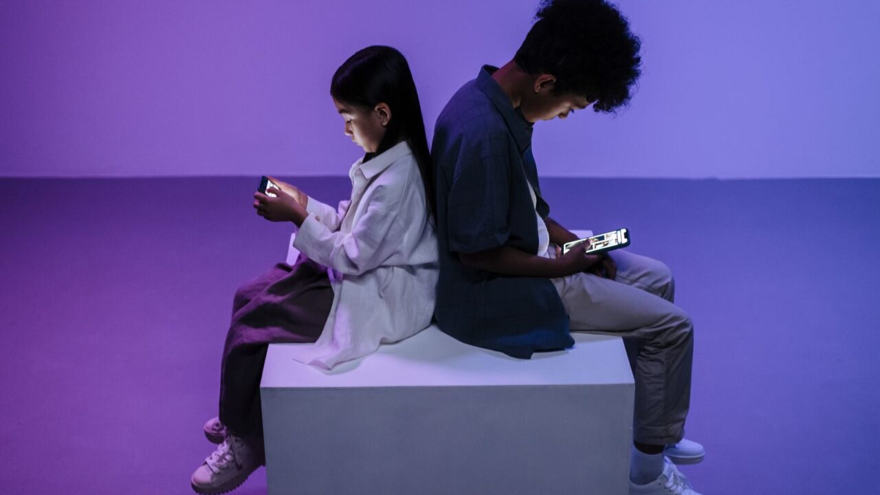 Two kids sitting back-to-back focused on smartphones, lit by moody lighting.