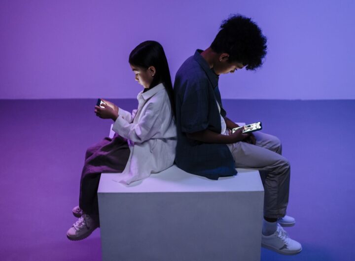 Two kids sitting back-to-back focused on smartphones, lit by moody lighting.