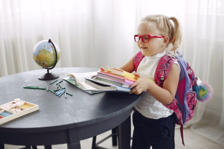 Little girl wearing casual clothes and glasses smiling while preparing books for homework at home during weekend and looking away