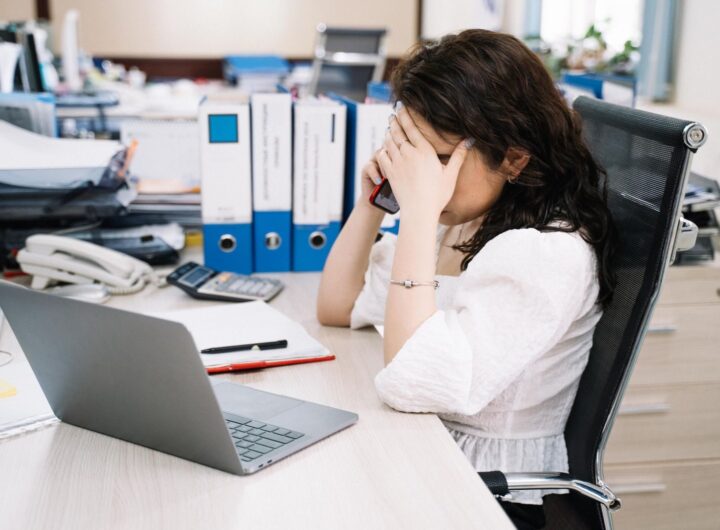 A woman in an office sitting at a desk, visibly stressed, using a phone. Workplace stress concept.