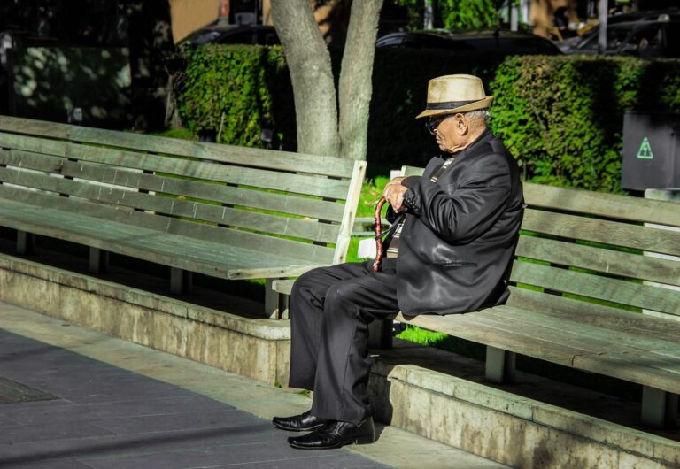 Senior man in a suit and hat resting on a bench outdoors, holding a cane in daylight.