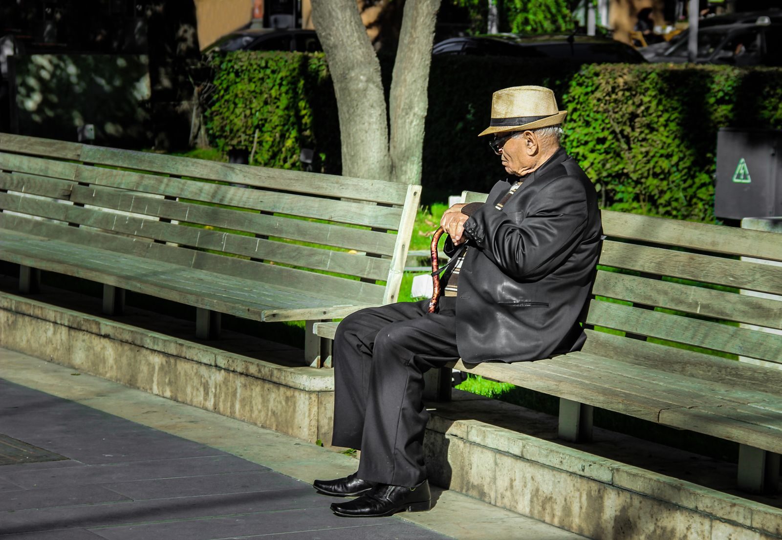 Senior man in a suit and hat resting on a bench outdoors, holding a cane in daylight.